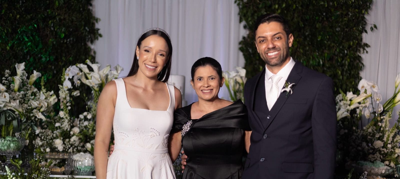 Weiri Borges entre uma convidada de vestido branco e o noivo, em cenário com flores brancas.