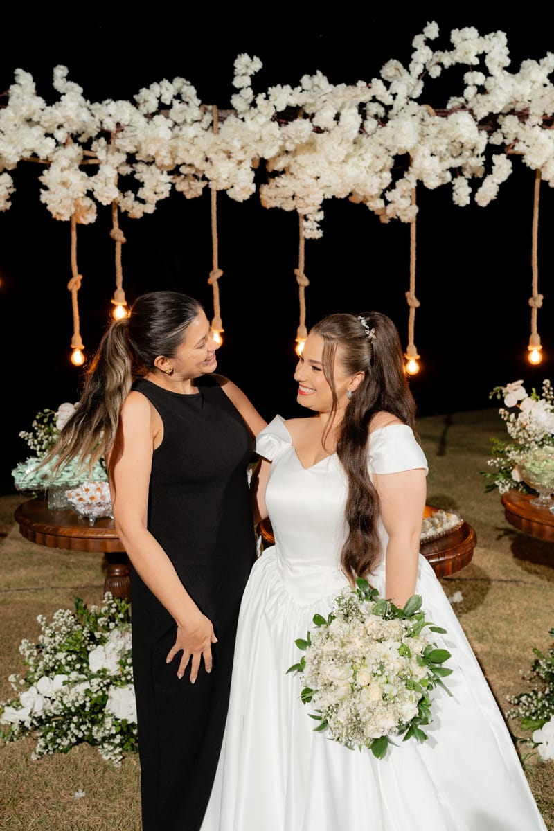 Weiri Borges e a noiva sorrindo uma para a outra sob arranjo de flores brancas.