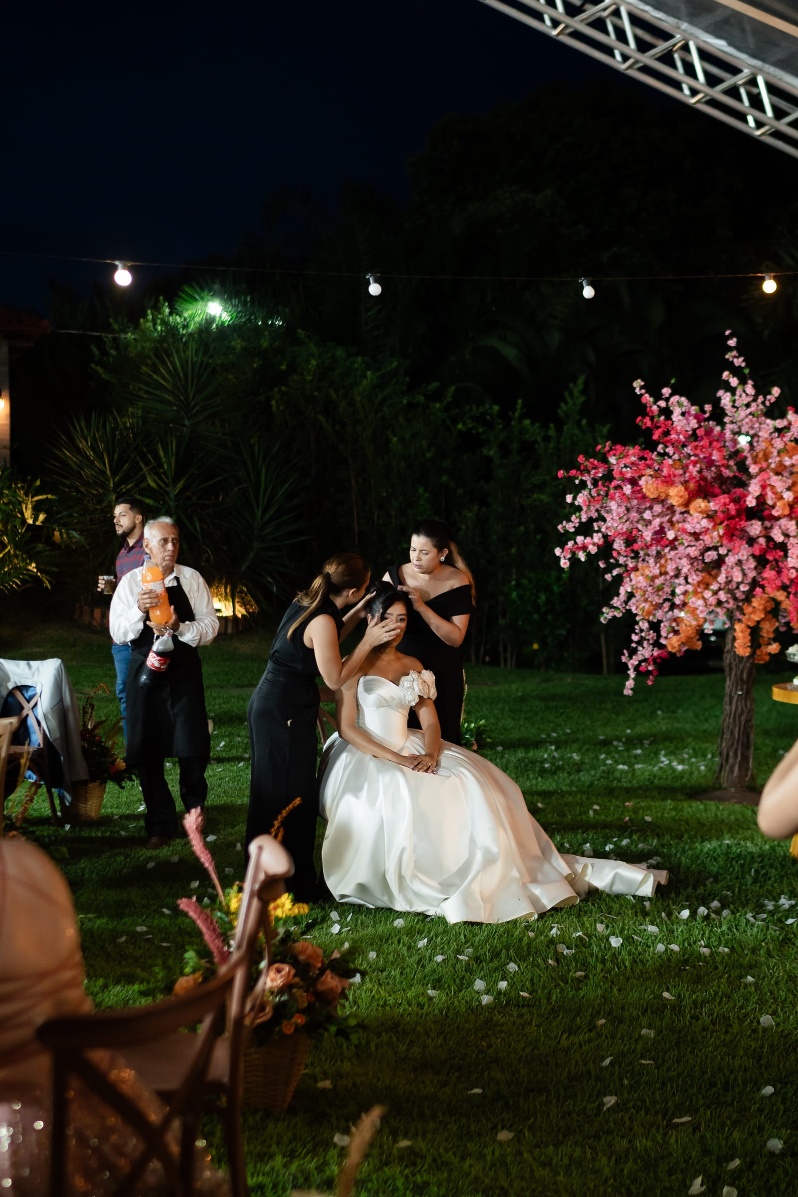 Equipe de Weiri Borges cuidando da noiva sentada no gramado, à noite, entre luzes e flores.